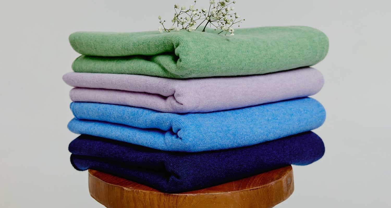 Folded cashmere in green, lilac, blue and navy, stacked on a wooden stool