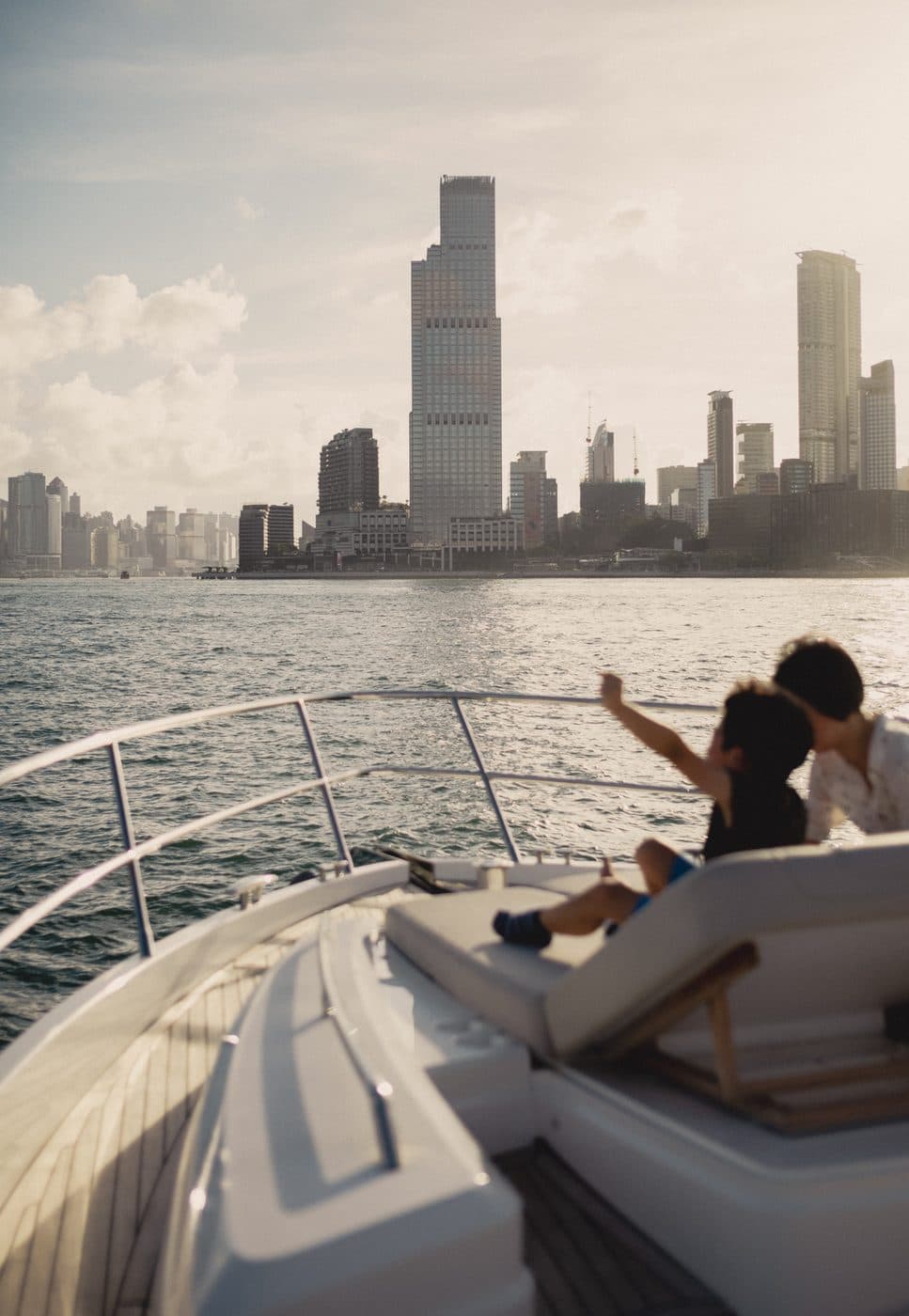 A child pointing from the bow of a boat on Victoria Harbour, Rosewood Hong Kong on the Kowloon waterfront behind